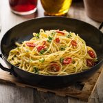 Su tavolo di legno, tagliere, una padella con Spaghetti aglio, olio e peperoncino , dietro una grattugia e un bicchiere di olio d'olive