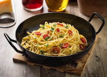 Su tavolo di legno, tagliere, una padella con Spaghetti aglio, olio e peperoncino , dietro una grattugia e un bicchiere di olio d'olive