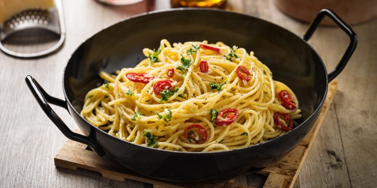 Su tavolo di legno, tagliere, una padella con Spaghetti aglio, olio e peperoncino , dietro una grattugia e un bicchiere di olio d'olive