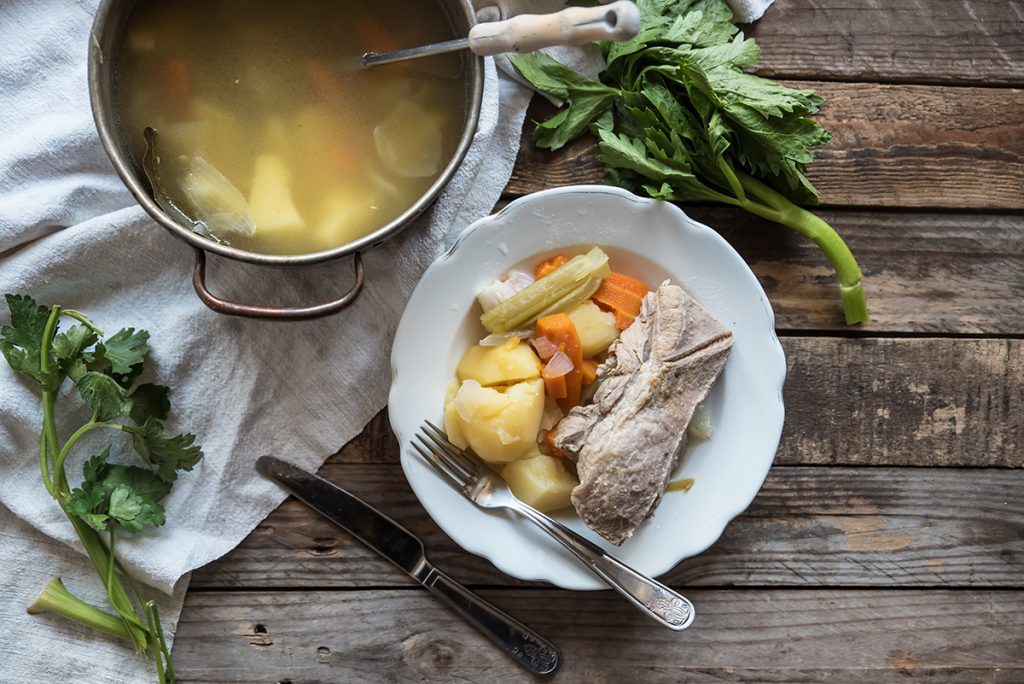 Su tavolo di legno, piatto bianco con carne, patate lesse e carote e su canovaccio bianco una pentola con bollito di carne