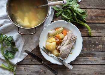 Su tavolo di legno, piatto bianco con carne, patate lesse e carote e su canovaccio bianco una pentola con bollito di carne