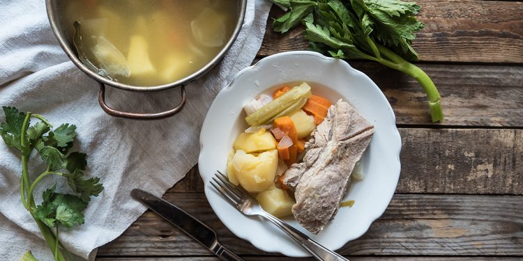 Su tavolo di legno, piatto bianco con carne, patate lesse e carote e su canovaccio bianco una pentola con bollito di carne