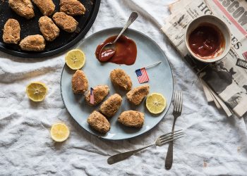 su un tavolo con tovaglia grigia piatto azzurro con nuggets di poll, un cucchiaino con ketchup e una piccola bandiera americana. Limoni tagliati nel piatto e attorni. Sulla destra foglie di giornali e un contenitore con Ketchup