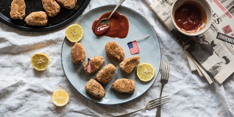 su un tavolo con tovaglia grigia piatto azzurro con nuggets di poll, un cucchiaino con ketchup e una piccola bandiera americana. Limoni tagliati nel piatto e attorni. Sulla destra foglie di giornali e un contenitore con Ketchup