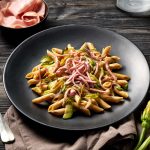 su tavolo di legno scuro, piatto nero con dentro pasta con mortadella e zucchine, accanto un piattino con mortadella e in basso a destra 2 fiori di zucchina accanto alla forchetta