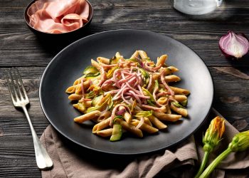 su tavolo di legno scuro, piatto nero con dentro pasta con mortadella e zucchine, accanto un piattino con mortadella e in basso a destra 2 fiori di zucchina accanto alla forchetta