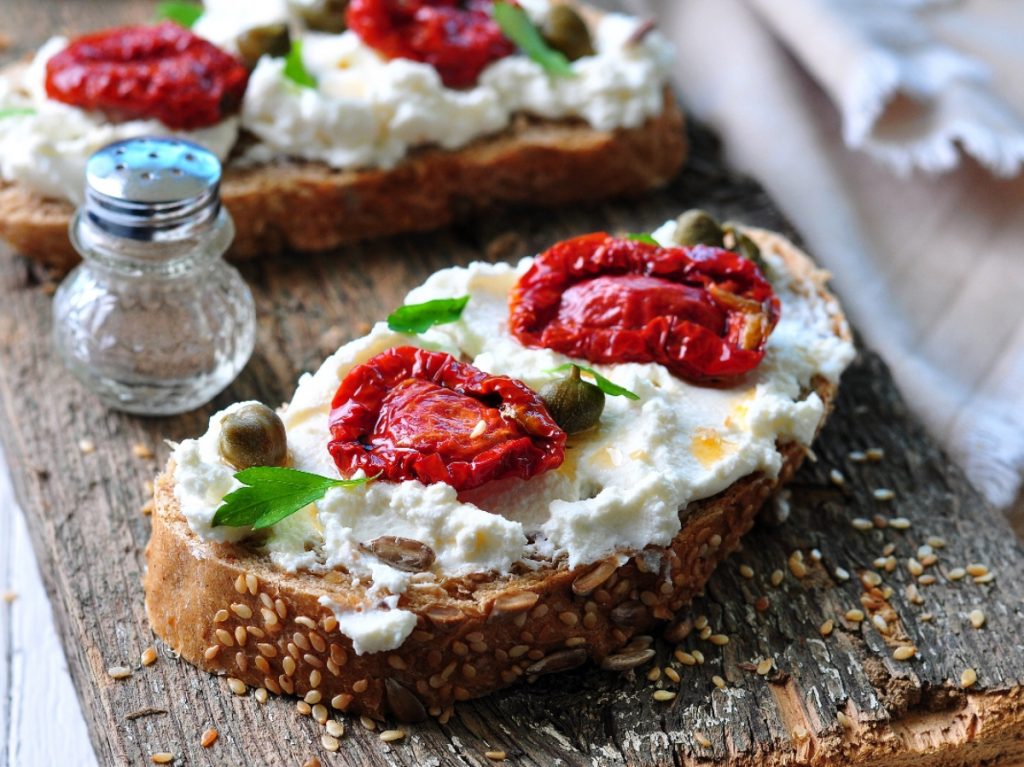 su tagliere di legno, primo piano di una bruschetta con ricotta e pomodori secchi, accanto pepe nero