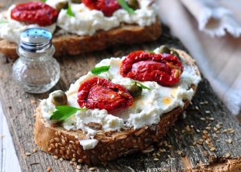 su tagliere di legno, primo piano di una bruschetta con ricotta e pomodori secchi, accanto pepe nero