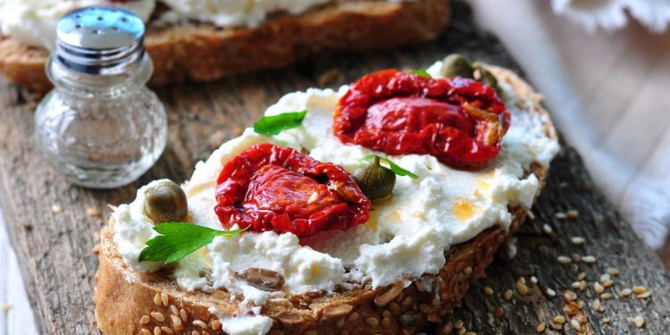 su tagliere di legno, primo piano di una bruschetta con ricotta e pomodori secchi, accanto pepe nero