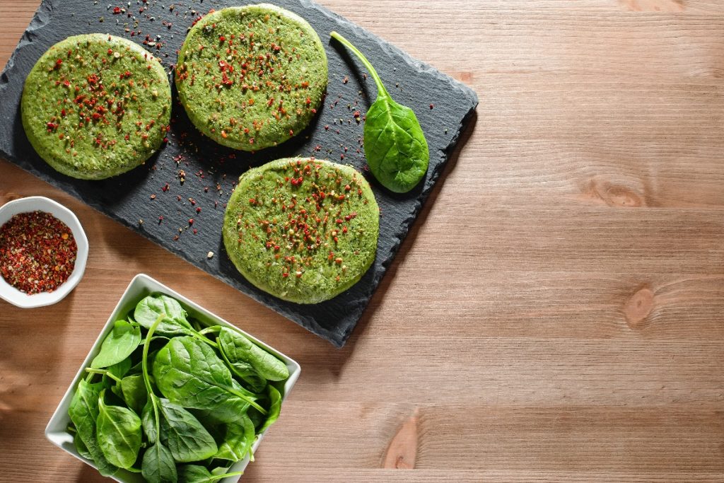 su tavolo di legno sotto piatto di ardesia con 3 burger di spinaci, un piccolo contenitore di peperoncino e a destra contenitore con spinaci freschi