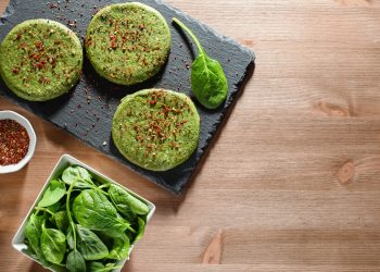 su tavolo di legno sotto piatto di ardesia con 3 burger di spinaci, un piccolo contenitore di peperoncino e a destra contenitore con spinaci freschi