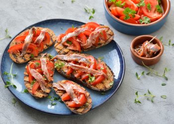 su tavolo di marmo, piatto blu con bruschette di pomodoro e alici . A destra contenitore con alici e dietro contenitore con pomodori