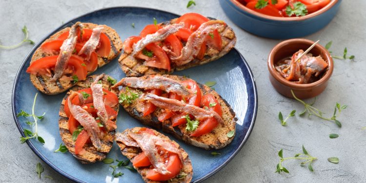 su tavolo di marmo, piatto blu con bruschette di pomodoro e alici . A destra contenitore con alici e dietro contenitore con pomodori