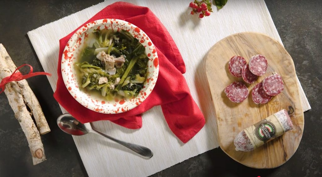 su tavolo nero tagliere bianco con un contenitore con minestra maritata con salamini sulla destra; tagliere di legno con un salame cacciatore e sulla sinistra 2 rami di legno con fiocco rosso