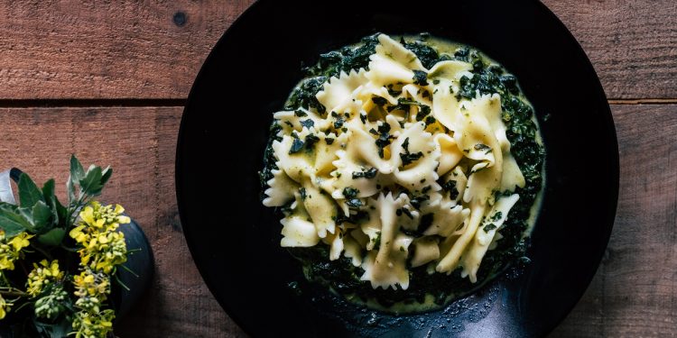 su tavolo di legno, piatto nero con pasta con spinaci freschi. A sinistra una pianta con fiori