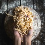 Su piano di lavoro di legno scuro, 2 mani che prendono tagliatelle fatte in casa. Sotto tagliere di legno pieno di farina