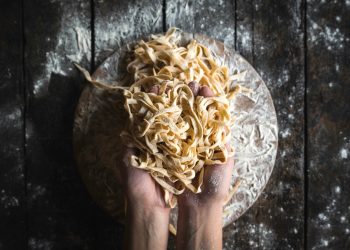 Su piano di lavoro di legno scuro, 2 mani che prendono tagliatelle fatte in casa. Sotto tagliere di legno pieno di farina