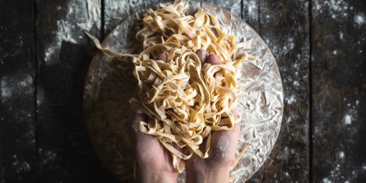 Su piano di lavoro di legno scuro, 2 mani che prendono tagliatelle fatte in casa. Sotto tagliere di legno pieno di farina