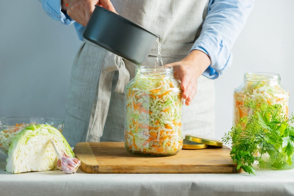 donna con grembiule che prepara in casa crauti versa acqua nel contenitore pieno, sulla sinistra cavolo cappuccio tagliato