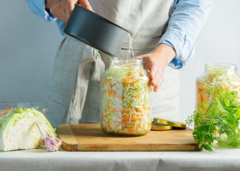 donna con grembiule che prepara in casa crauti versa acqua nel contenitore pieno, sulla sinistra cavolo cappuccio tagliato