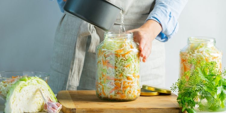 donna con grembiule che prepara in casa crauti versa acqua nel contenitore pieno, sulla sinistra cavolo cappuccio tagliato