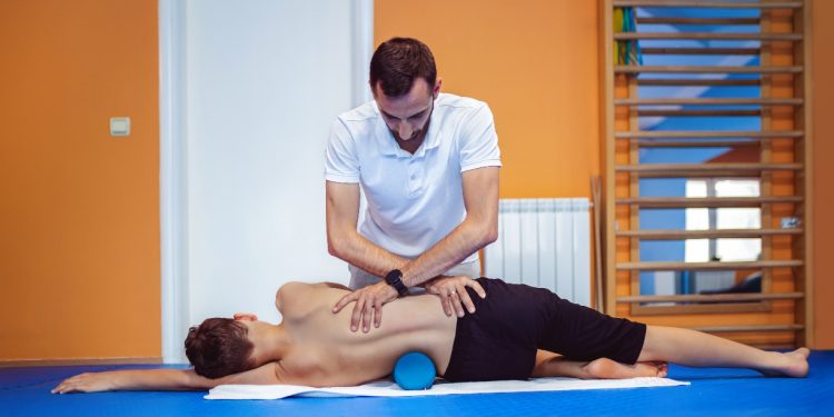 Nello studio di kinesiologia, il terapista pratica posturale sulla schiena di un ragazzo che è disteso di profilo e ha sotto un foam roller