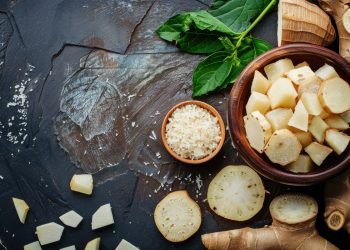 su tavolo nero, ciotola con manioca tagliata a fette con altre fette della radice sparse sul tavolo e erbe aromatiche