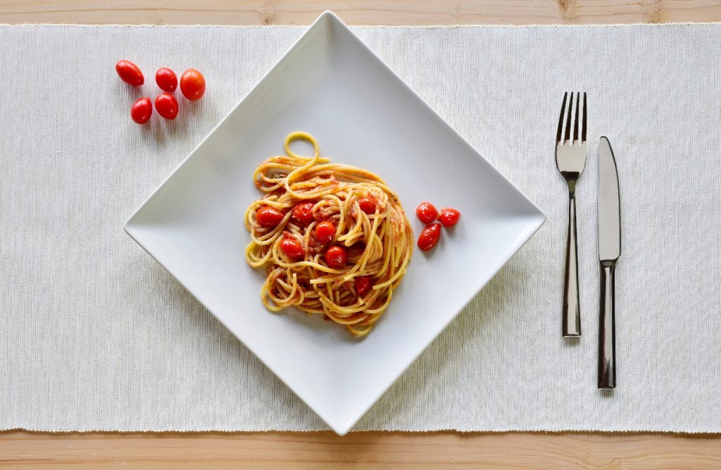 su tavolo di legno sottopiatto bianco di stoffa con un un piatto quadrato pieno di pasta con pomodorini freschi. Accanto al piatto 5 pomodorini e a destra forchette e coltello