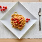 su tavolo di legno sottopiatto bianco di stoffa con un un piatto quadrato pieno di pasta con pomodorini freschi. Accanto al piatto 5 pomodorini e a destra forchette e coltello