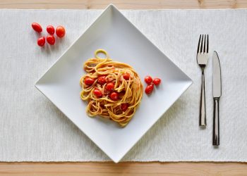 su tavolo di legno sottopiatto bianco di stoffa con un un piatto quadrato pieno di pasta con pomodorini freschi. Accanto al piatto 5 pomodorini e a destra forchette e coltello