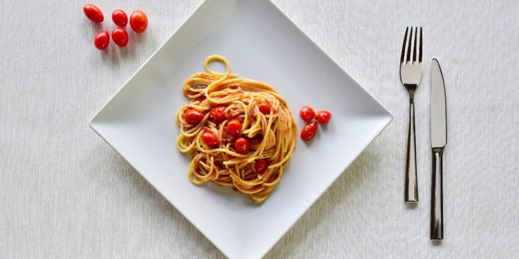 su tavolo di legno sottopiatto bianco di stoffa con un un piatto quadrato pieno di pasta con pomodorini freschi. Accanto al piatto 5 pomodorini e a destra forchette e coltello