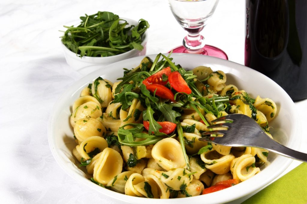 su tavolo con tovaglia bianca, un piatto bianco con pasta con rucola e pomodoro (orecchiette), vicino un bicchiere d'acqua e un contenitore di rucola