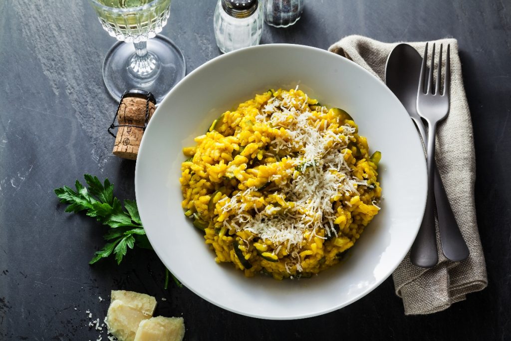 Su piano di cottura nero, piatto bianco con un risotto alla zafferano con riso Carnaroli, attorno al piatto, scaglie di parmiggiano, tappo del vino, saliera e pepiera, forchetta e coltello