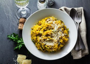 Su piano di cottura nero, piatto bianco con un risotto alla zafferano con riso Carnaroli, attorno al piatto, scaglie di parmiggiano, tappo del vino, saliera e pepiera, forchetta e coltello