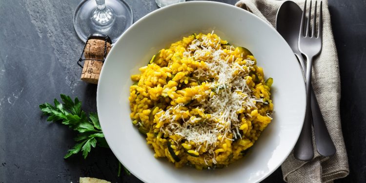 Su piano di cottura nero, piatto bianco con un risotto alla zafferano con riso Carnaroli, attorno al piatto, scaglie di parmiggiano, tappo del vino, saliera e pepiera, forchetta e coltello