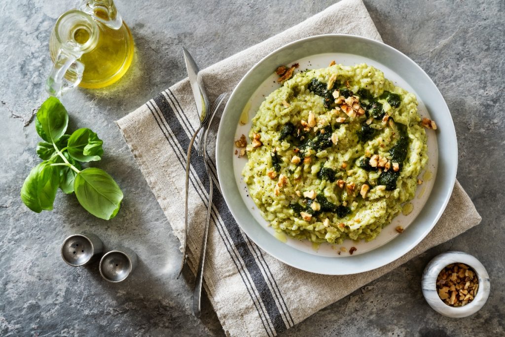 su tovaglia grigia, canovaccio con sopra un piatto che contiene risotto con zucchine e frutta secca con forchette e coltello, a sinistra basilico e contenitore di olio EVO