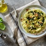 su tovaglia grigia, canovaccio con sopra un piatto che contiene risotto con zucchine e frutta secca con forchette e coltello, a sinistra basilico e contenitore di olio EVO