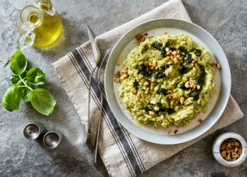 su tovaglia grigia, canovaccio con sopra un piatto che contiene risotto con zucchine e frutta secca con forchette e coltello, a sinistra basilico e contenitore di olio EVO