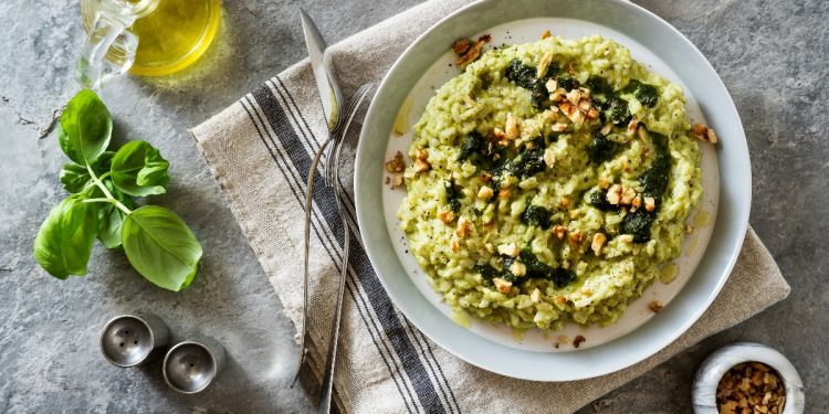 su tovaglia grigia, canovaccio con sopra un piatto che contiene risotto con zucchine e frutta secca con forchette e coltello, a sinistra basilico e contenitore di olio EVO