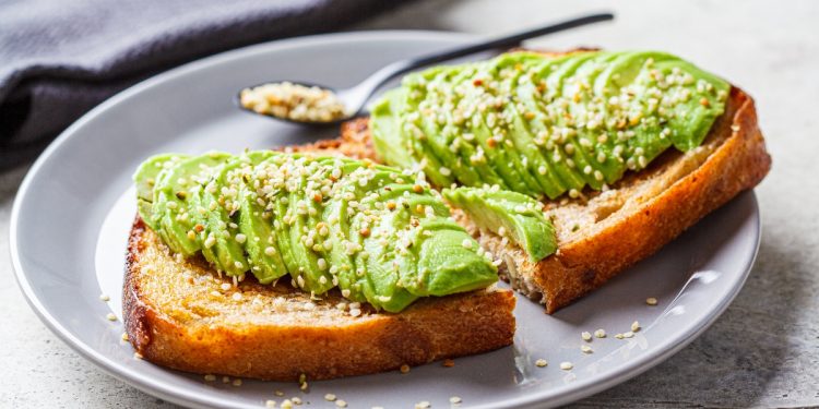 su tavolo di legno grigio, toast con avocado e semi di canapa e un cucchiaino. Dietro tovagliolo grigio