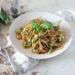 su tavolo di marmo, piatto fondo con spaghetti alla Nerano con zucchine e basilico, sulla sinistra cucchiaio e forchetta e dietro il piatto una zucchina tagliata in 2 e un contenitore pepe