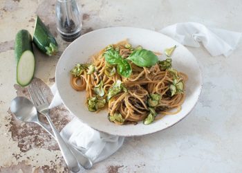 su tavolo di marmo, piatto fondo con spaghetti alla Nerano con zucchine e basilico, sulla sinistra cucchiaio e forchetta e dietro il piatto una zucchina tagliata in 2 e un contenitore pepe