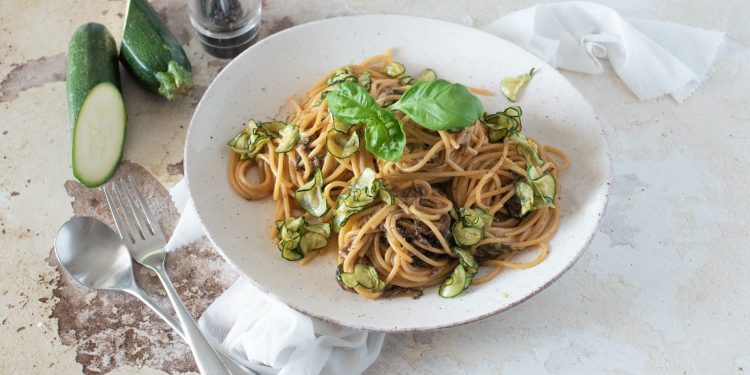 su tavolo di marmo, piatto fondo con spaghetti alla Nerano con zucchine e basilico, sulla sinistra cucchiaio e forchetta e dietro il piatto una zucchina tagliata in 2 e un contenitore pepe