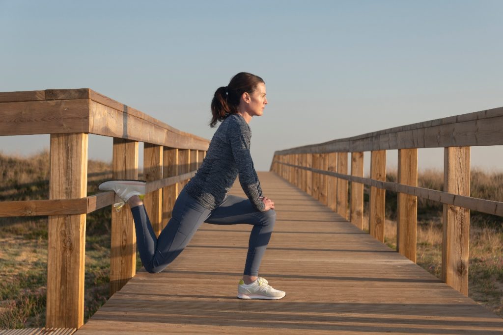 Donna con completo fitness grigio e scarpe bianche da fitness esegue su un ponte stretching gambe in affondo con il piede sul ponte di legno per allungare muscolatura