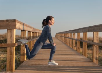 Donna con completo fitness grigio e scarpe bianche da fitness esegue su un ponte stretching gambe in affondo con il piede sul ponte di legno per allungare muscolatura