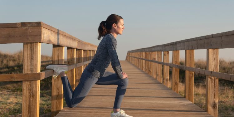 Donna con completo fitness grigio e scarpe bianche da fitness esegue su un ponte stretching gambe in affondo con il piede sul ponte di legno per allungare muscolatura