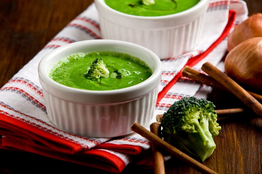 su tavolo di legno, canovaccio bianco e rosso piegato con 2 ciotole piccole con vellutata di broccoli con bacchette cinesi e accanto un pezzo di broccolo