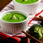 su tavolo di legno, canovaccio bianco e rosso piegato con 2 ciotole piccole con vellutata di broccoli con bacchette cinesi e accanto un pezzo di broccolo