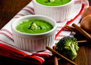 su tavolo di legno, canovaccio bianco e rosso piegato con 2 ciotole piccole con vellutata di broccoli con bacchette cinesi e accanto un pezzo di broccolo