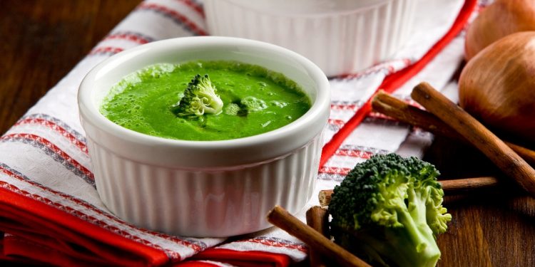 su tavolo di legno, canovaccio bianco e rosso piegato con 2 ciotole piccole con vellutata di broccoli con bacchette cinesi e accanto un pezzo di broccolo